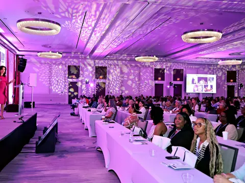 Speaker presenting on stage to a large audience during a conference in a ballroom illuminated with purple lighting.