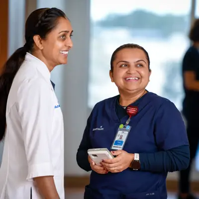 Two smiling AdventHealth employees, one in a white coat and the other in blue scrubs, engage in conversation.