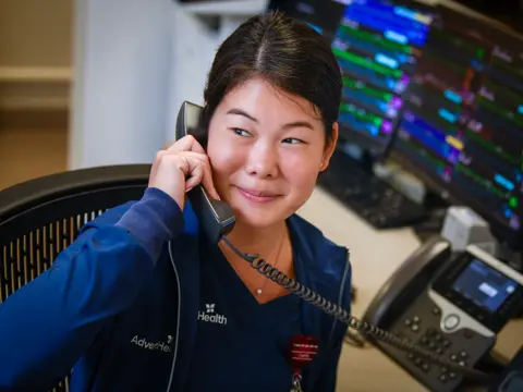 A smiling healthcare worker in a blue uniform talks on the phone in a medical office.