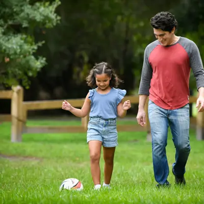 A father and daughter walking together on a grassy field with a soccer ball nearby.