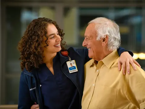 A smiling nurse with curly hair embraces an elderly man, both wearing AdventHealth badges.