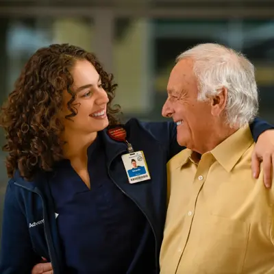 A smiling nurse with curly hair embraces an elderly man, both wearing AdventHealth badges.