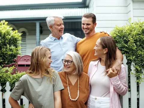 Five smiling adults of varying ages stand closely together outdoors in front of a house with white picket fence and garden, sharing a warm family moment