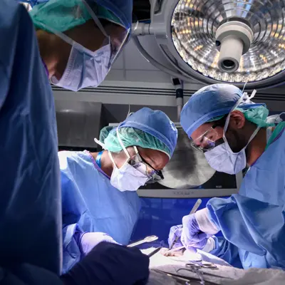 Surgeons in blue scrubs and masks perform surgery under a bright surgical light.