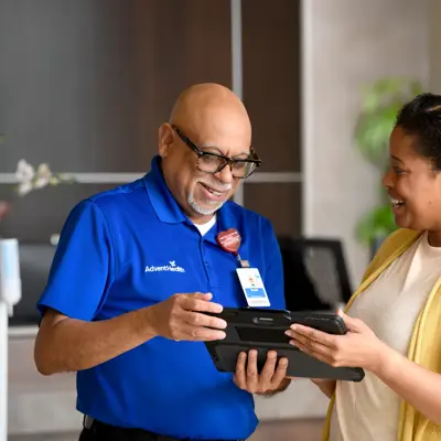 A happy AdventHealth employee shows a guest something on a tablet while they stand at the Guest Service desk.