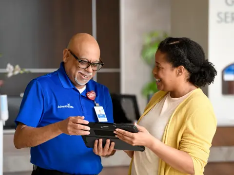 A happy AdventHealth employee shows a guest something on a tablet while they stand at the Guest Service desk.