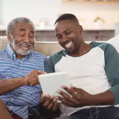 Two men sitting on a couch, smiling and looking at a tablet.