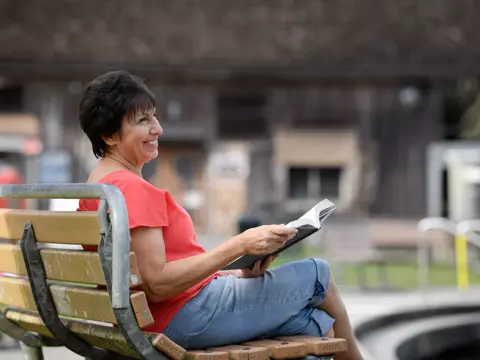 A smiling woman in a red top and blue jeans sits on a bench reading a book.