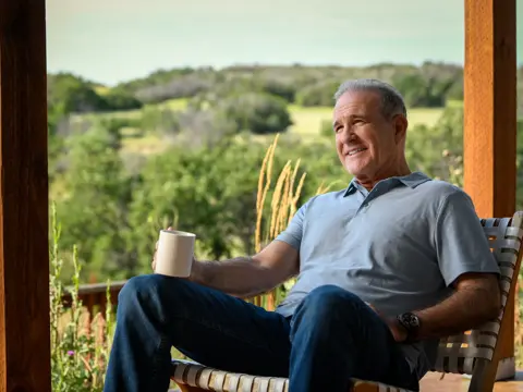 Smiling man in a gray shirt and blue jeans sits on a porch holding a coffee mug.