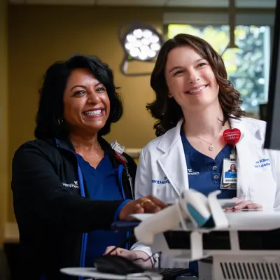 Two smiling healthcare professionals stand by a computer in a medical setting.