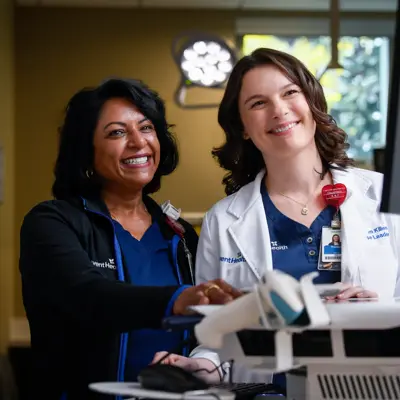 Two smiling healthcare professionals stand by a computer in a medical setting.