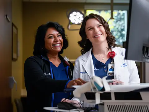 Two smiling healthcare professionals stand by a computer in a medical setting.