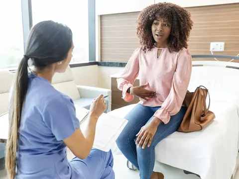 Woman in pink blouse gesturing with her hand while describing stomach pain to a female doctor in blue scrubs seated across from her in a modern medical consultation room