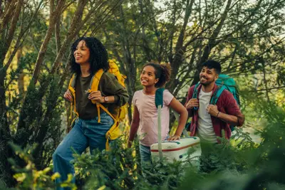 Smiling family hiking through a forest, carrying backpacks and a cooler, enjoying nature together on a sunny day.