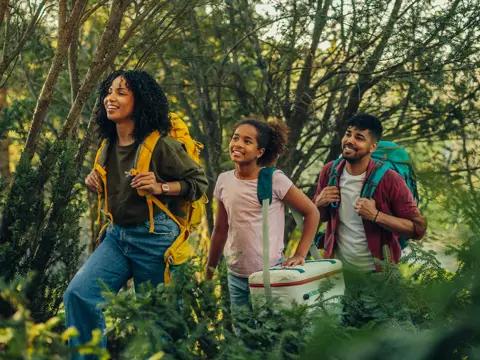 Smiling family hiking through a forest, carrying backpacks and a cooler, enjoying nature together on a sunny day.