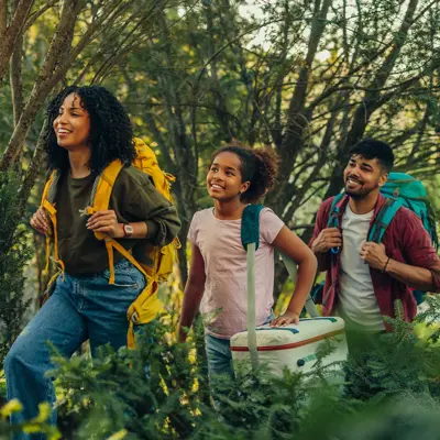 Smiling family hiking through a forest, carrying backpacks and a cooler, enjoying nature together on a sunny day.