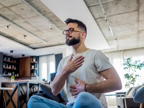 Young man with glasses and beard wearing a white t-shirt practicing breathing exercises while seated in a modern home interior with kitchen and plants visible in the background