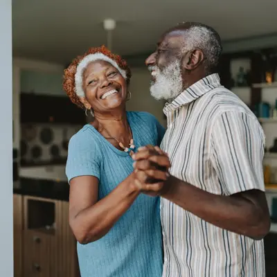Smiling older couple holding hands and dancing together in a bright living room.