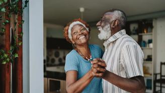 Smiling older couple holding hands and dancing together in a bright living room.