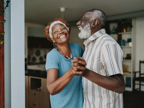 Smiling older couple holding hands and dancing together in a bright living room.