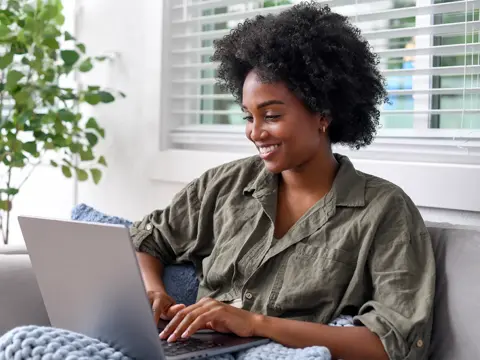 Woman with curly hair smiling while using a laptop on a gray sofa in a bright living room with plants and windows