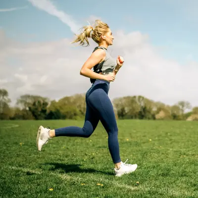 Woman in athletic wear runs across a grassy field under a blue sky with light clouds.
