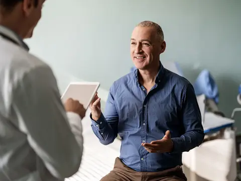 Smiling man gestures while speaking with a person in a white coat holding a tablet in an exam room.