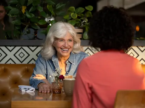 A smiling woman with white hair sits at a table in a restaurant, facing another person.