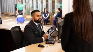 A smiling man in a suit sits at a desk with a computer, assisting a woman.