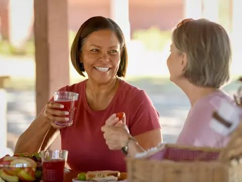Two women smiling and enjoying a picnic with fruit and drinks.