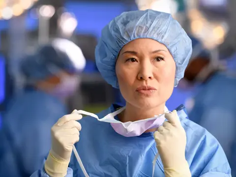A female surgeon in scrubs and a surgical cap holds a mask in an operating room.