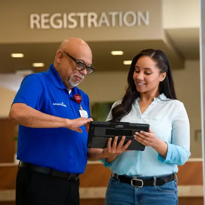 A smiling woman holds a tablet as a man points to the screen in front of a registration sign.