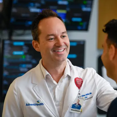 Healthcare provider in white coat smiling while speaking with a patient in a clinical room with medical monitoring equipment in the background