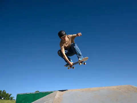 A skateboarder in a brown shirt and blue jeans performs a trick in mid-air above a ramp.