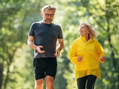 Two people walk on a wooded path, one in a yellow jacket, talking while moving forward.