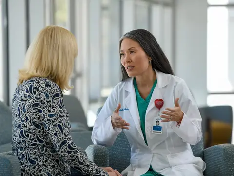 A doctor in a white coat and green scrubs talks with a patient in a waiting room.