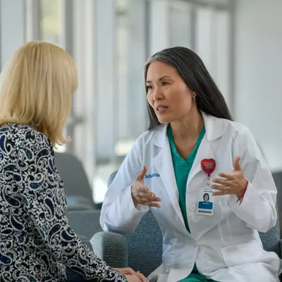A doctor in a white coat and green scrubs talks with a patient in a waiting room.