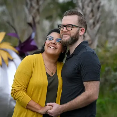 A smiling couple stands close together in front of a large, colorful metal flower sculpture.