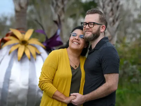 A smiling couple stands close together in front of a large, colorful metal flower sculpture.