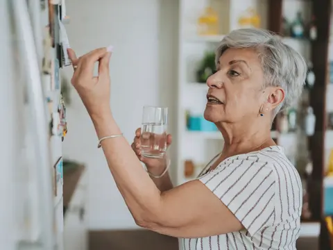 Senior woman with short gray hair holding a glass of water while checking a wall calendar in her home kitchen