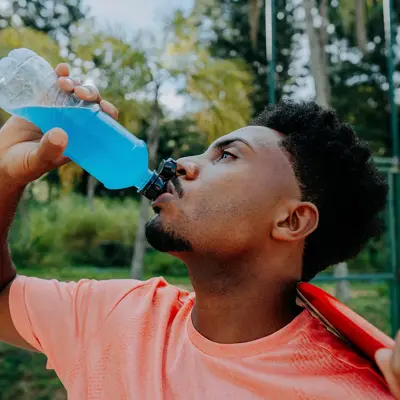 Man drinking a blue sports drink from a bottle while outdoors after exercising, with another person holding a massage gun against his neck.