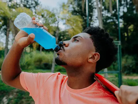 Man drinking a blue sports drink from a bottle while outdoors after exercising, with another person holding a massage gun against his neck.