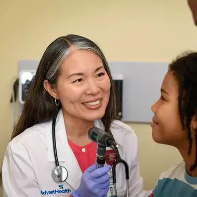 Doctor uses an otoscope to examine a child’s ear while an adult stands close beside the child.