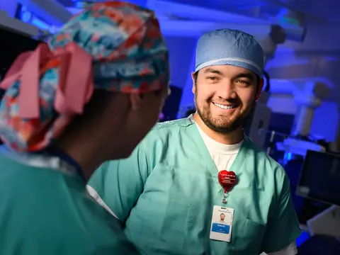 Smiling male surgeon in scrubs and cap stands in hospital room with female colleague.