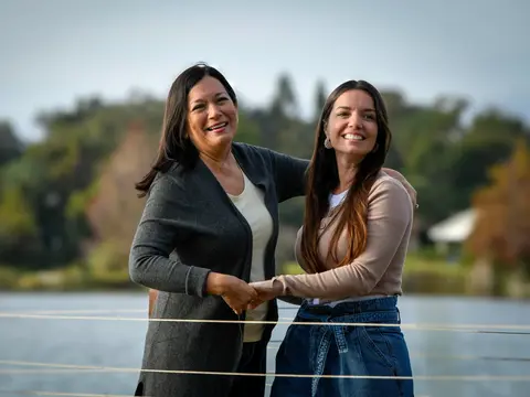Two smiling Hispanic women standing together outdoors by a lake with trees in the background, one wearing a dark cardigan and the other in a beige top