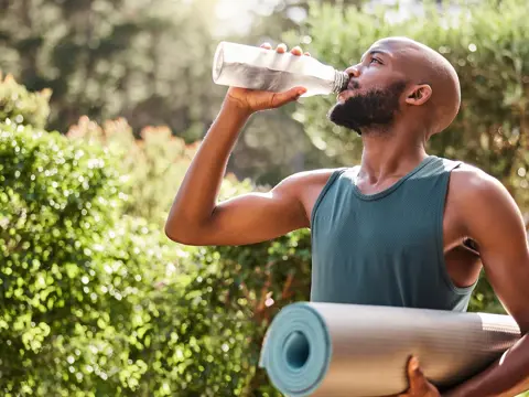Man in a teal tank top drinking water from a clear bottle while holding a rolled yoga mat outdoors in a sunny park setting