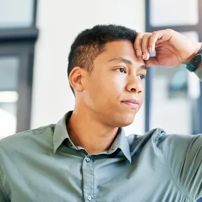 Man in green button-up shirt with hand on forehead appearing stressed or in pain while sitting in an office setting