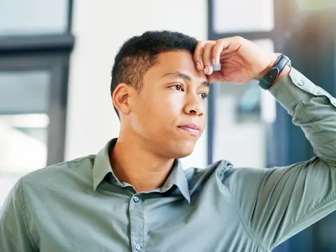 Man in green button-up shirt with hand on forehead appearing stressed or in pain while sitting in an office setting