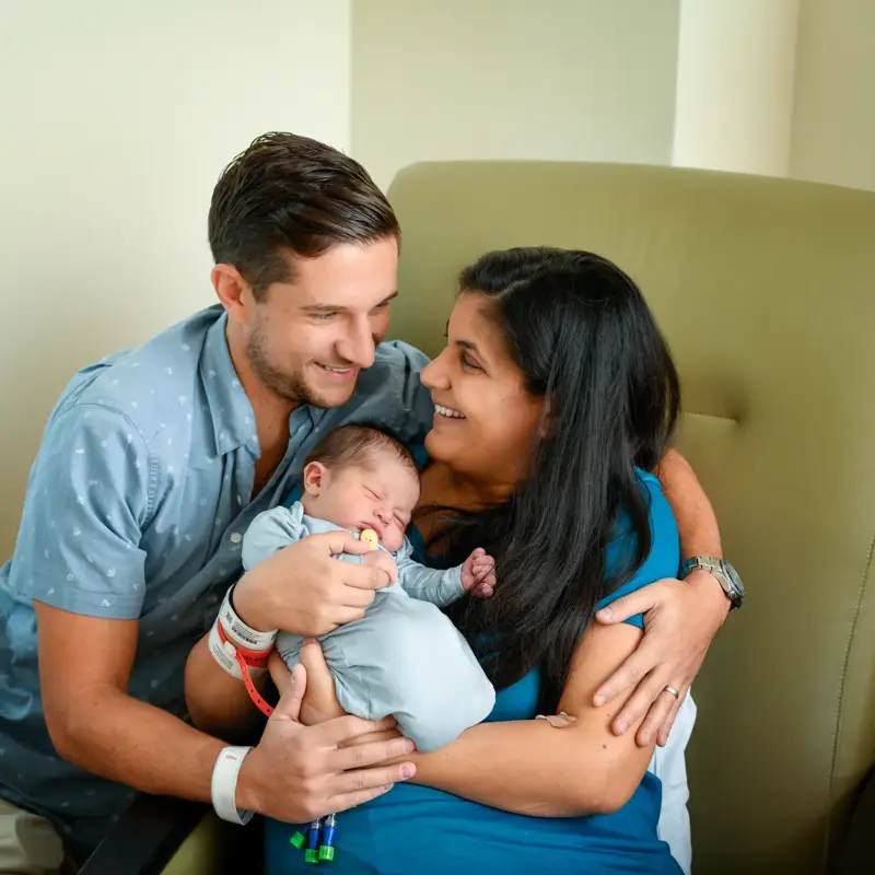 New parents embrace their sleeping newborn while sitting together on a hospital chair, sharing a joyful moment.