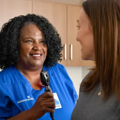 A smiling AdventHealth nurse in blue scrubs holds an otoscope while talking to a patient.